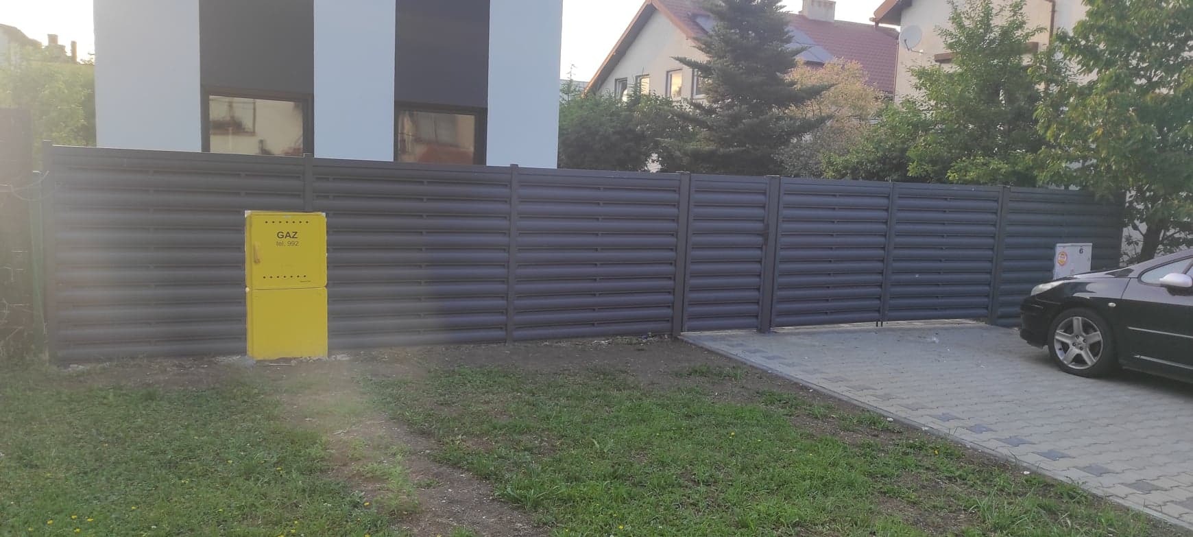 Nowoczesne, poziome ogrodzenie z szarych paneli blaszanych, przylegające do budynku z żółtą skrzynką gazową i podjazdem z kostki brukowej, częściowo zasłonięte przez zaparkowany samochód.