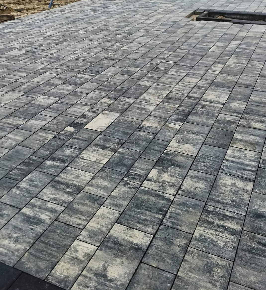 Ułożona nawierzchnia z prostokątnej kostki w odcieniach szarości i bieli, z widocznym fragmentem przygotowanego wykopu pod kolejne elementy.