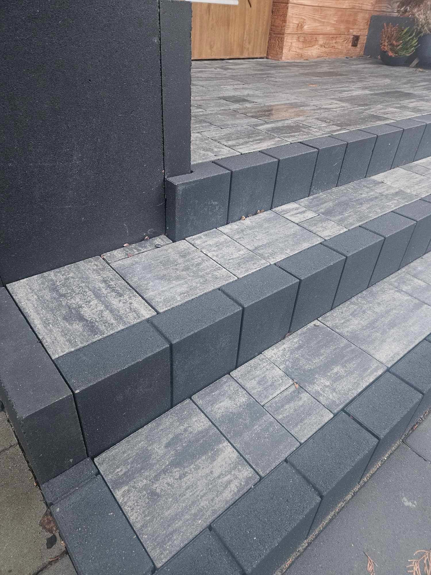 Schody wejściowe wyłożone dwukolorową kostką brukową w odcieniach szarości i antracytu, prowadzące do drewnianego wejścia z donicami po bokach.