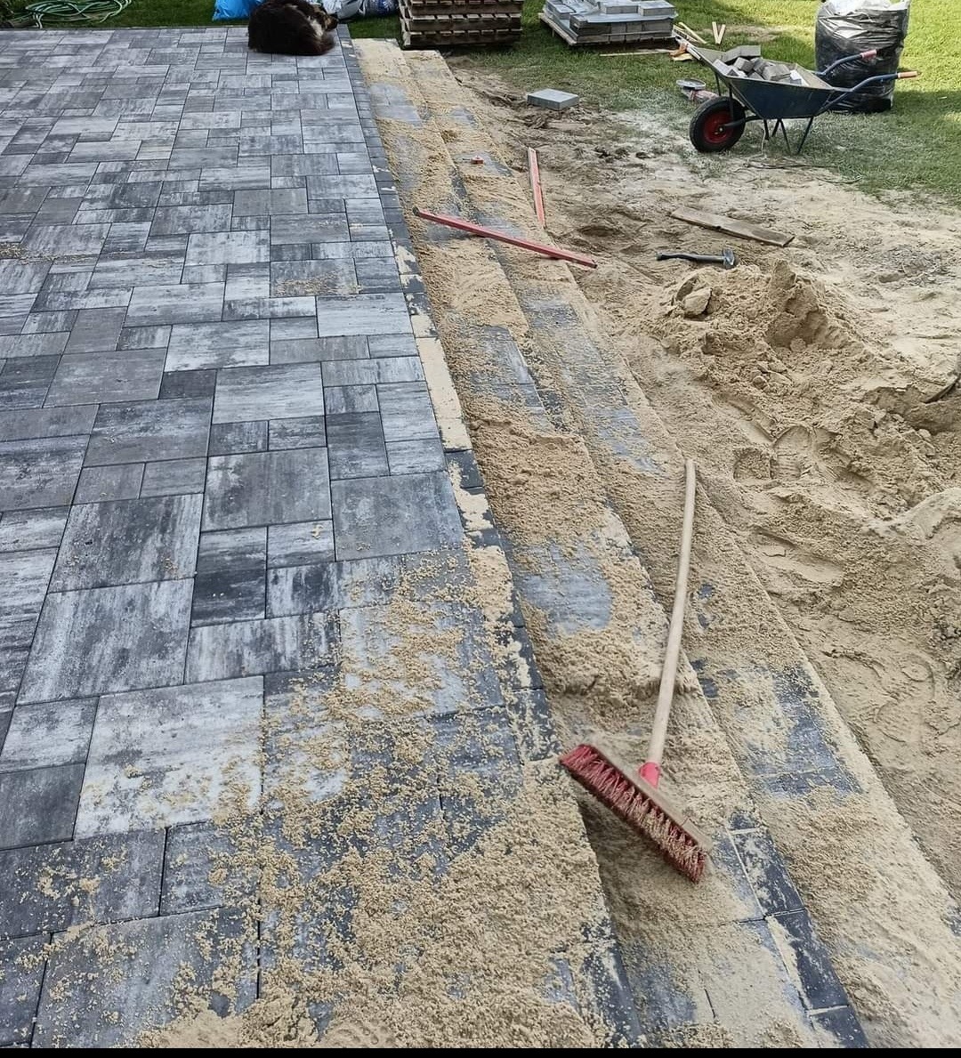 Fragment układanej szarej kostki brukowej z piaskiem, widoczny czerwony zamiatacz, łopata i taczka z kostką w tle.