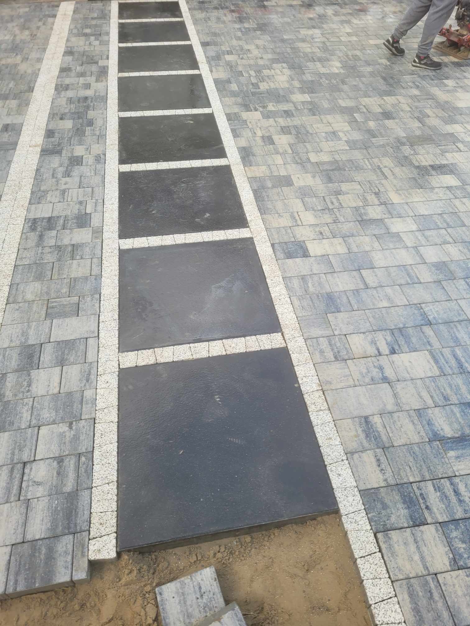 Niedokończona ścieżka z szarej kostki brukowej z wstawkami z dużych, czarnych płyt, obramowana jasną kostką; widoczne fragmenty piasku i nogi osoby w tle.