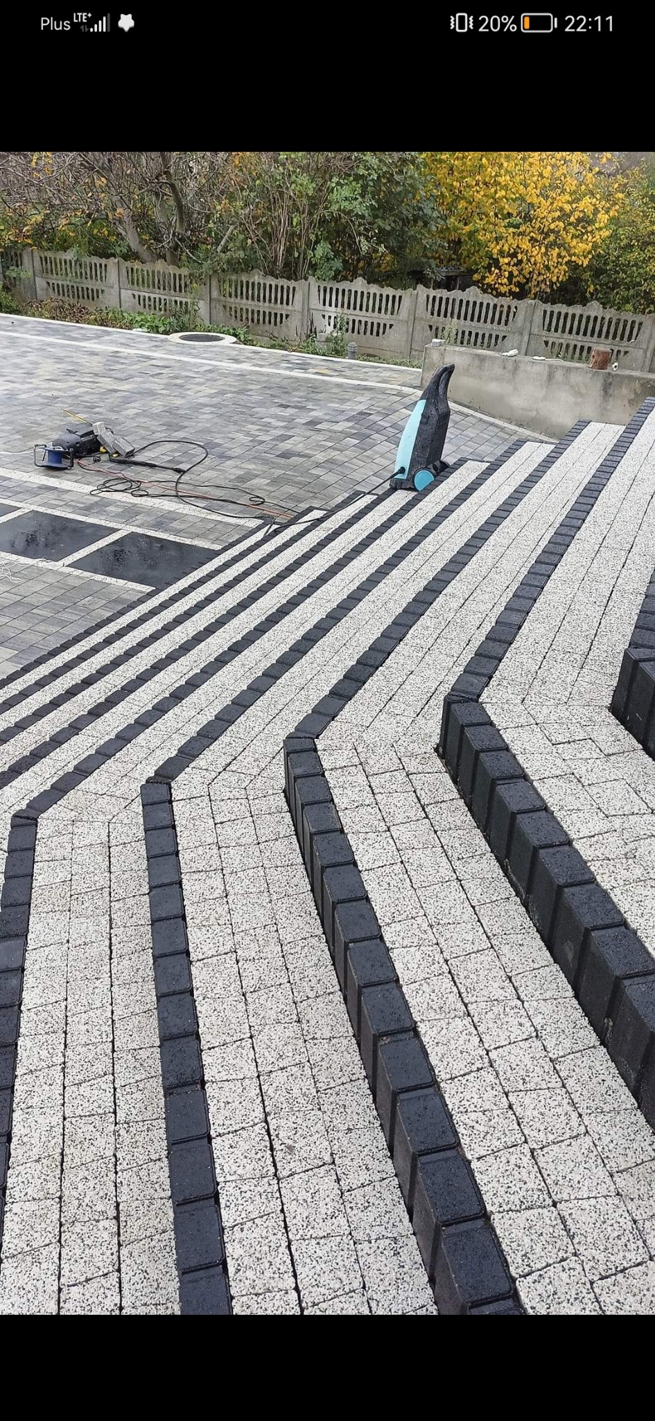 Ułożona kostka brukowa w kontrastujących kolorach szarości i czerni tworząca wzór na schodach zewnętrznych, w tle widoczny betonowy płot i zieleń, na kostce leży maszyna do czyszczenia kostki...