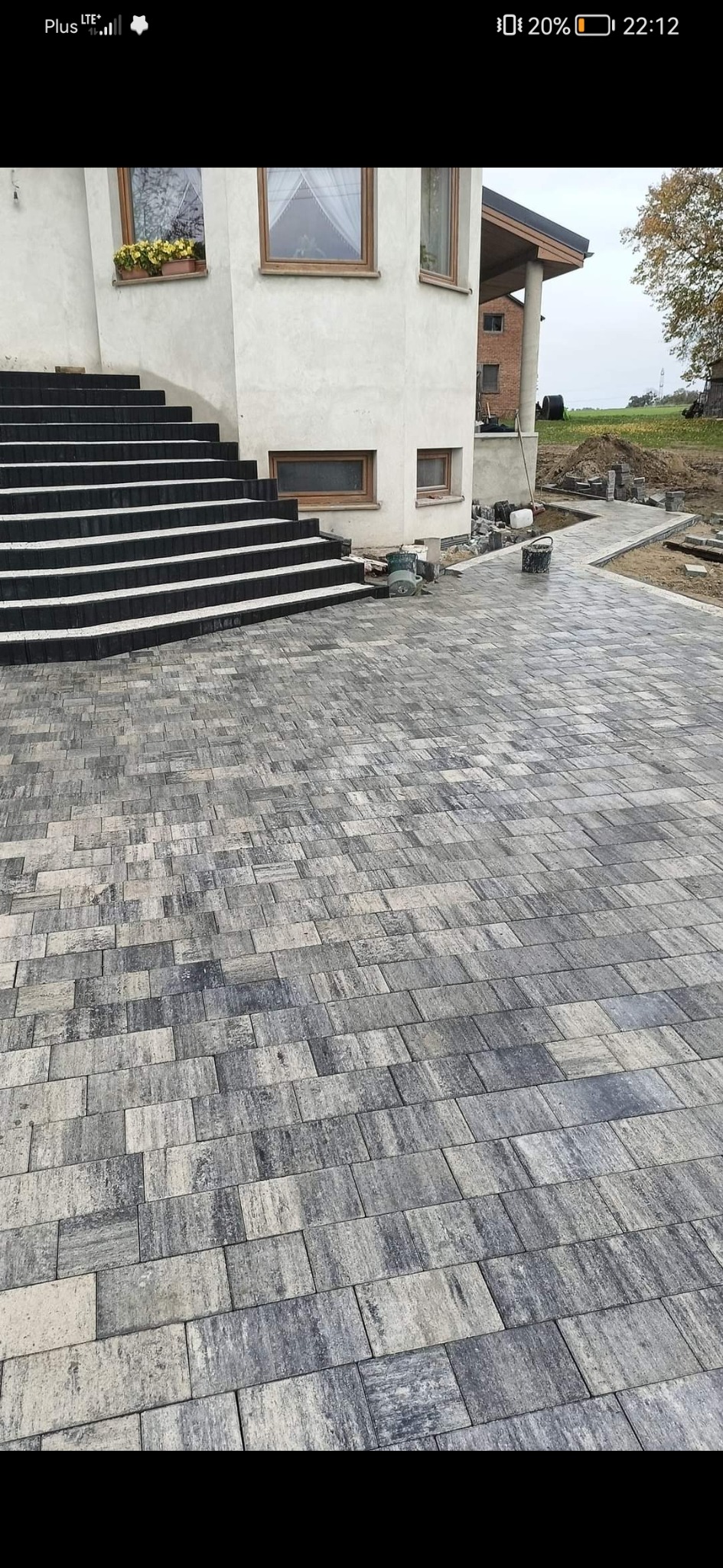 Ułożona szara kostka brukowa prowadząca od schodów domu do ścieżki w ogrodzie. Widoczne wiadro i narzędzia brukarskie.