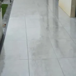 Jasnoszary taras wyłożony dużymi, kwadratowymi płytami. Widoczne fugi, ściana budynku w tle i fragment zieleni po lewej stronie.