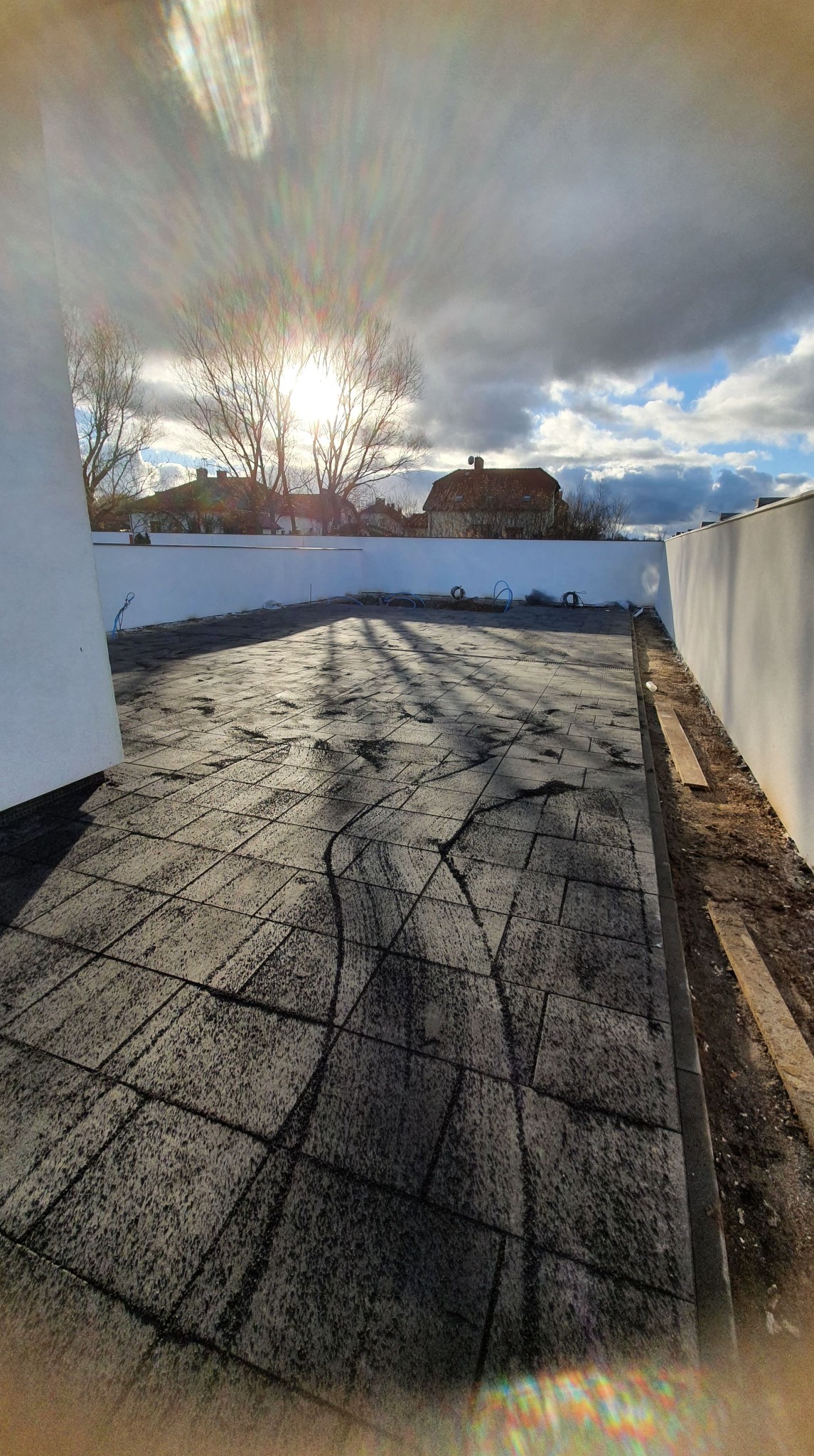 Ułożona, szara kostka brukowa na tarasie z białym murem w tle, oświetlona popołudniowym słońcem, z widocznymi cieniami drzew.