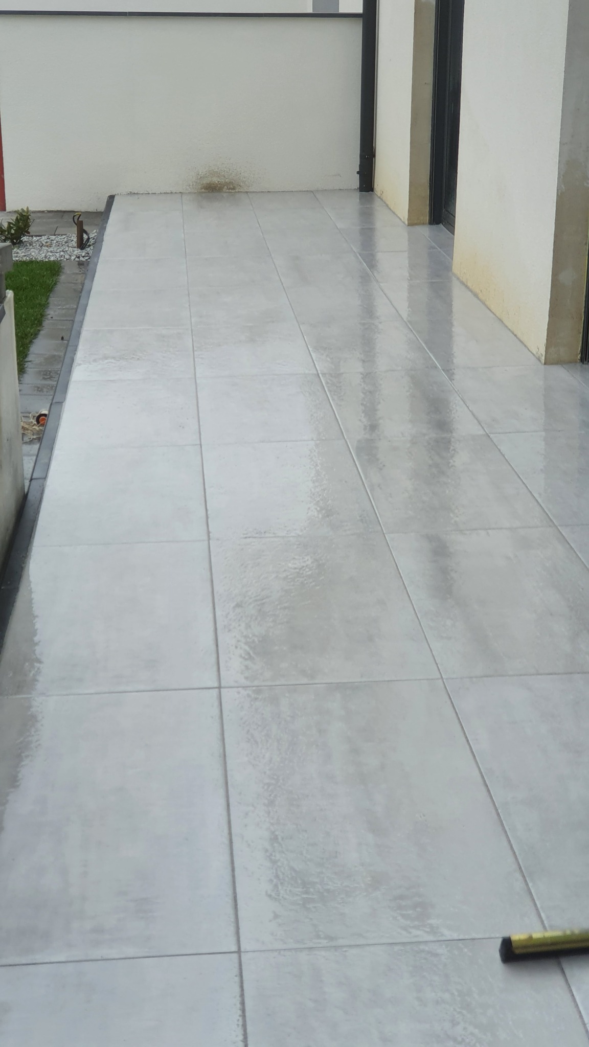 Jasnoszary taras wyłożony dużymi, kwadratowymi płytami. Widoczne fugi, ściana budynku w tle i fragment zieleni po lewej stronie.