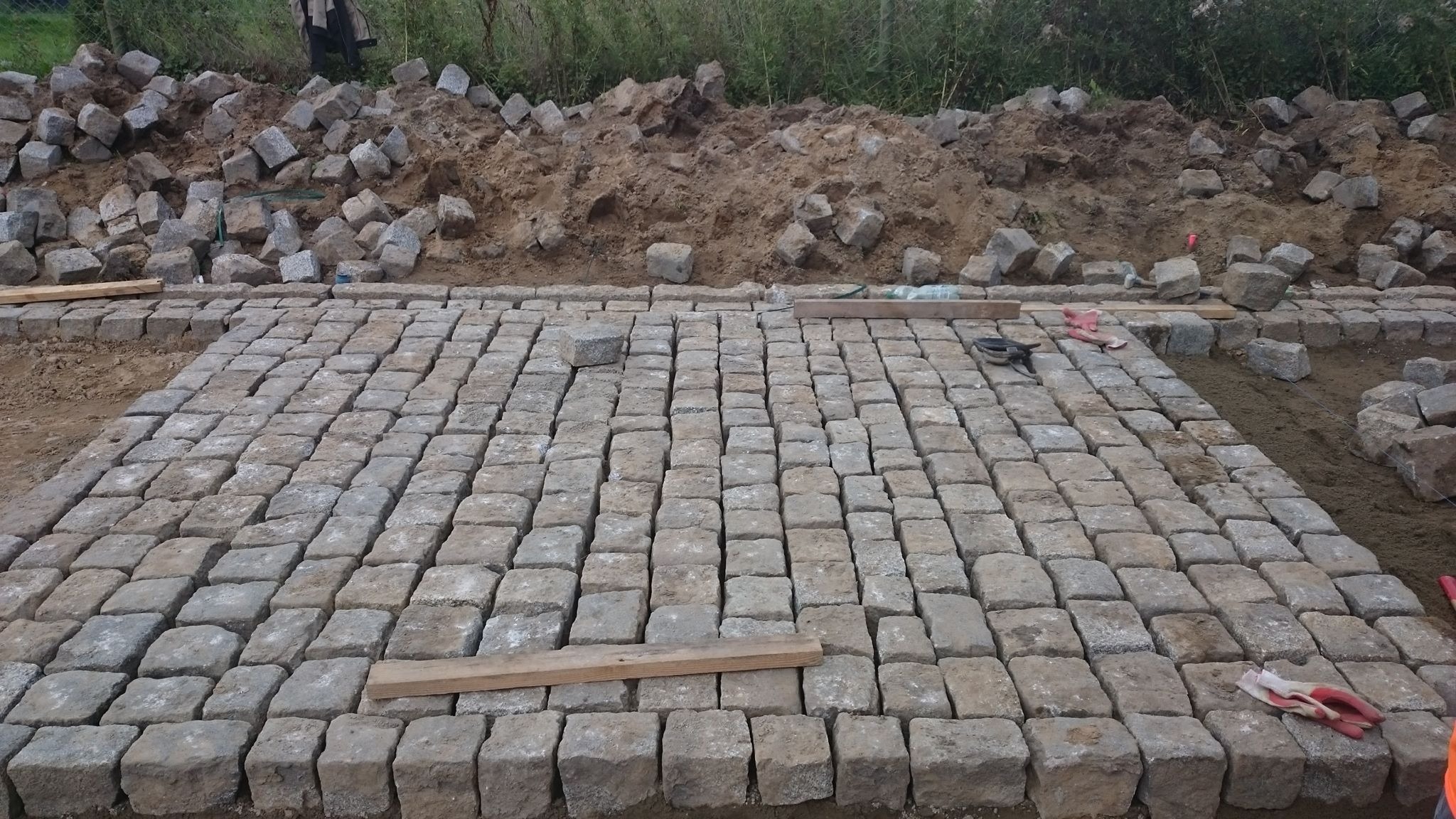 Fragment układanej powierzchni z kostki granitowej, widoczne narzędzia, rękawice robocze oraz pryzma kostki w tle, etap prac.