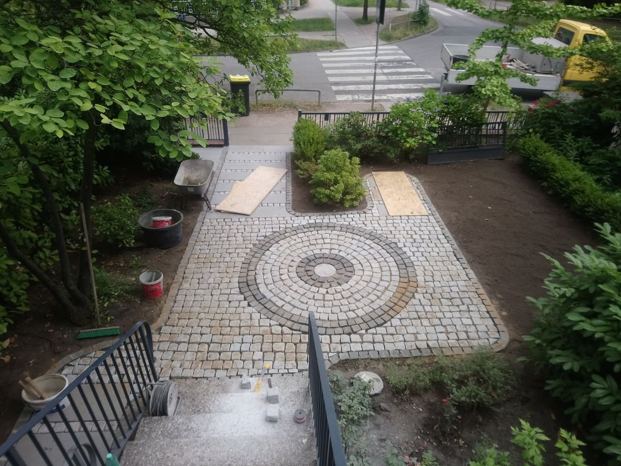 Widok z góry na ścieżkę z kostki granitowej ułożoną w okrągły wzór z jasnoszarego i beżowego granitu, w tle przygotowany teren pod trawnik i elementy zieleni. Na schodach widoczne narzędzia...