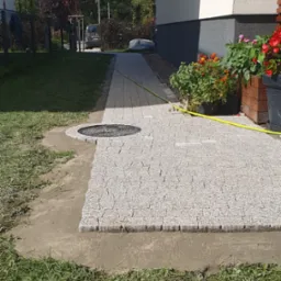 Układanie kostki granitowej na ścieżce obok domu, widoczny fragment trawnika i studzienka kanalizacyjna, donice z kwiatami przy ścianie.