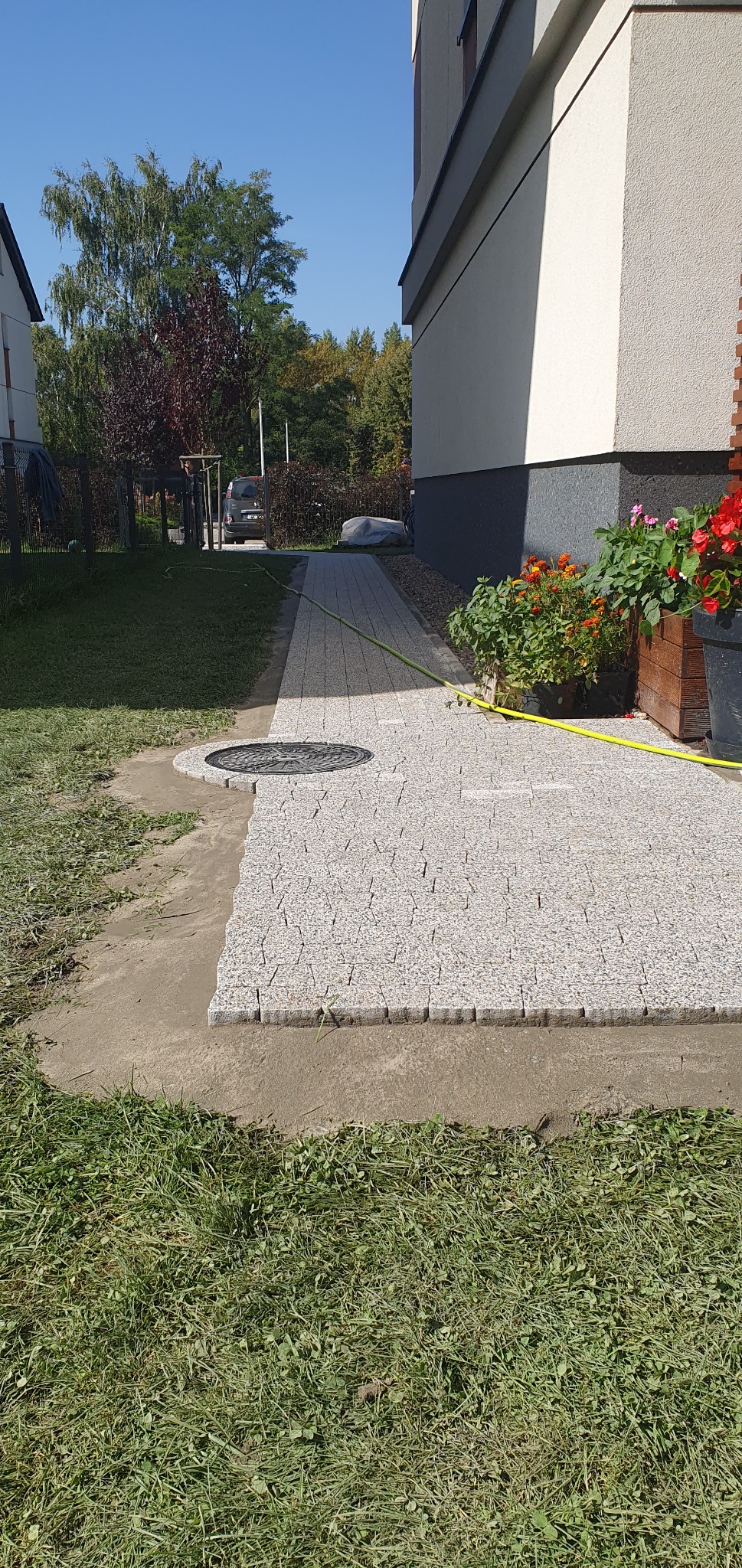 Układanie kostki granitowej na ścieżce obok domu, widoczny fragment trawnika i studzienka kanalizacyjna, donice z kwiatami przy ścianie.
