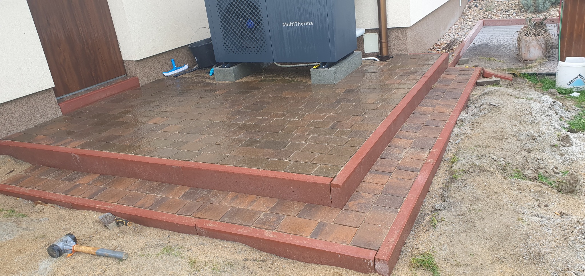 Brązowa kostka brukowa ułożona w schody wejściowe do domu, z widocznym agregatem MultiTherma i gumowym młotkiem na piasku.