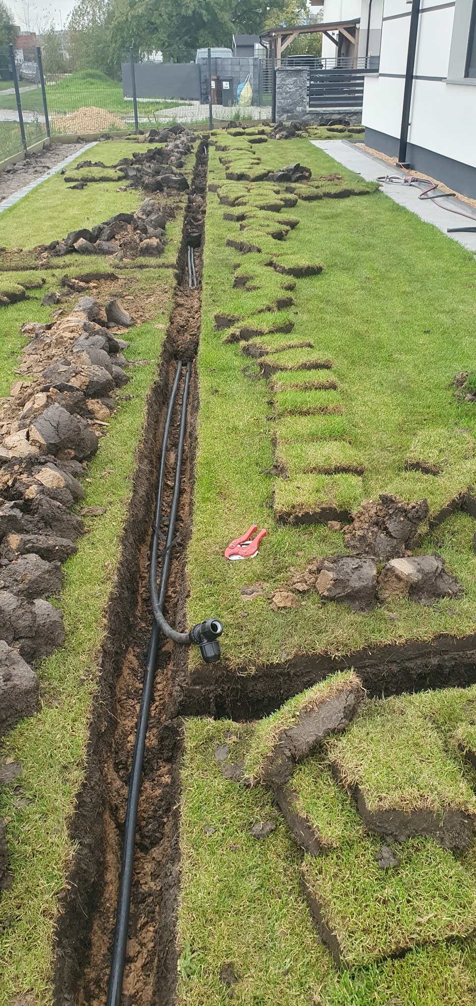 Wykopany rów w trawniku z ułożonymi rurami, wycięte kwadraty darni obok rowu, czerwone nożyce do rur leżą na trawie.