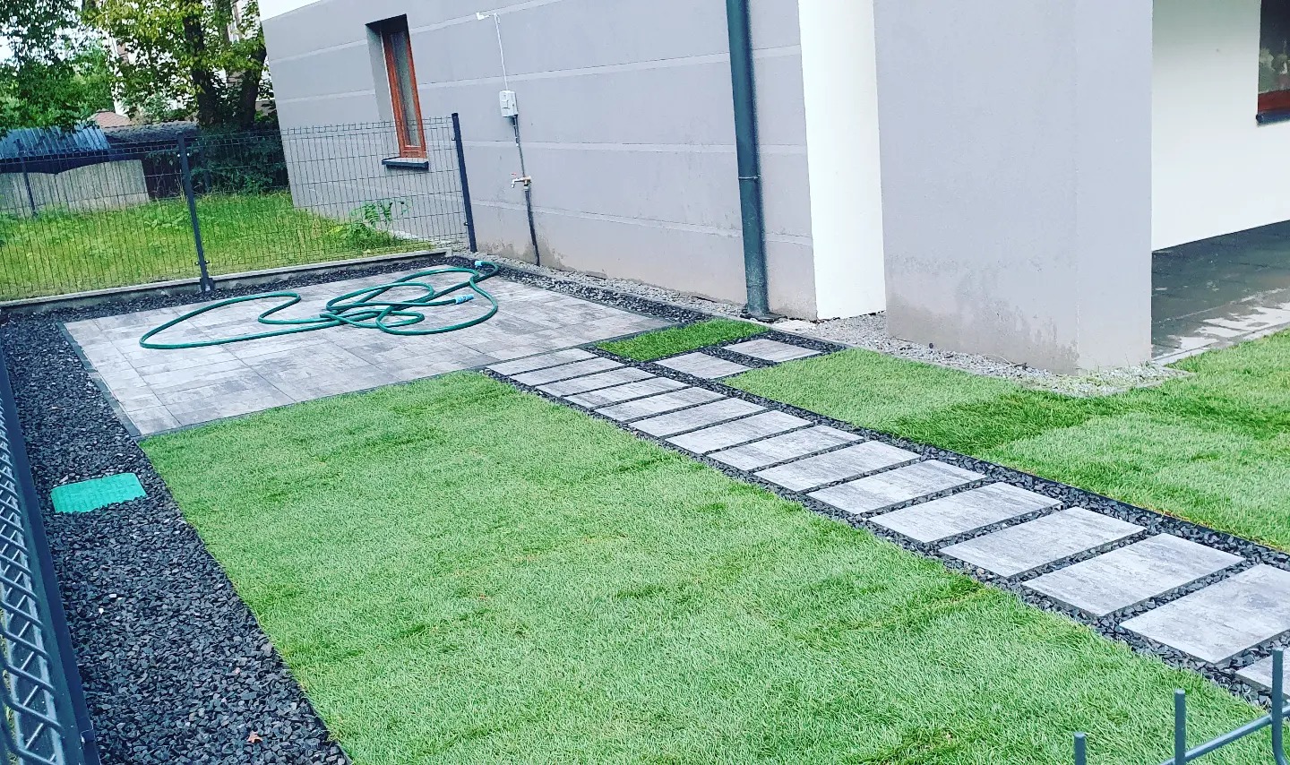 Ścieżka z kwadratowych płyt betonowych prowadząca przez trawnik, obrzeża z czarnego grysu, w tle budynek z szarą elewacją i zwinięty wąż ogrodowy.