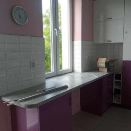 Konrad - Fragment kuchni w trakcie remontu: szare płytki nad blatem, fioletowe szafki, okno z widokiem na zieleń, widoczne narzędzia i materiały budowlane na blacie, oraz kartony na szafce.