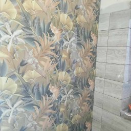 Konrad - Ściana z wzorzystą tapetą w motywy roślinne sąsiadująca z ułożonymi szarymi płytkami ceramicznymi w trakcie prac wykończeniowych.