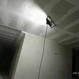 Konrad - Świeżo położona gładź na suficie podświetlona lampą budowlaną, widoczne ślady szlifowania, drabina aluminiowa w rogu pomieszczenia.