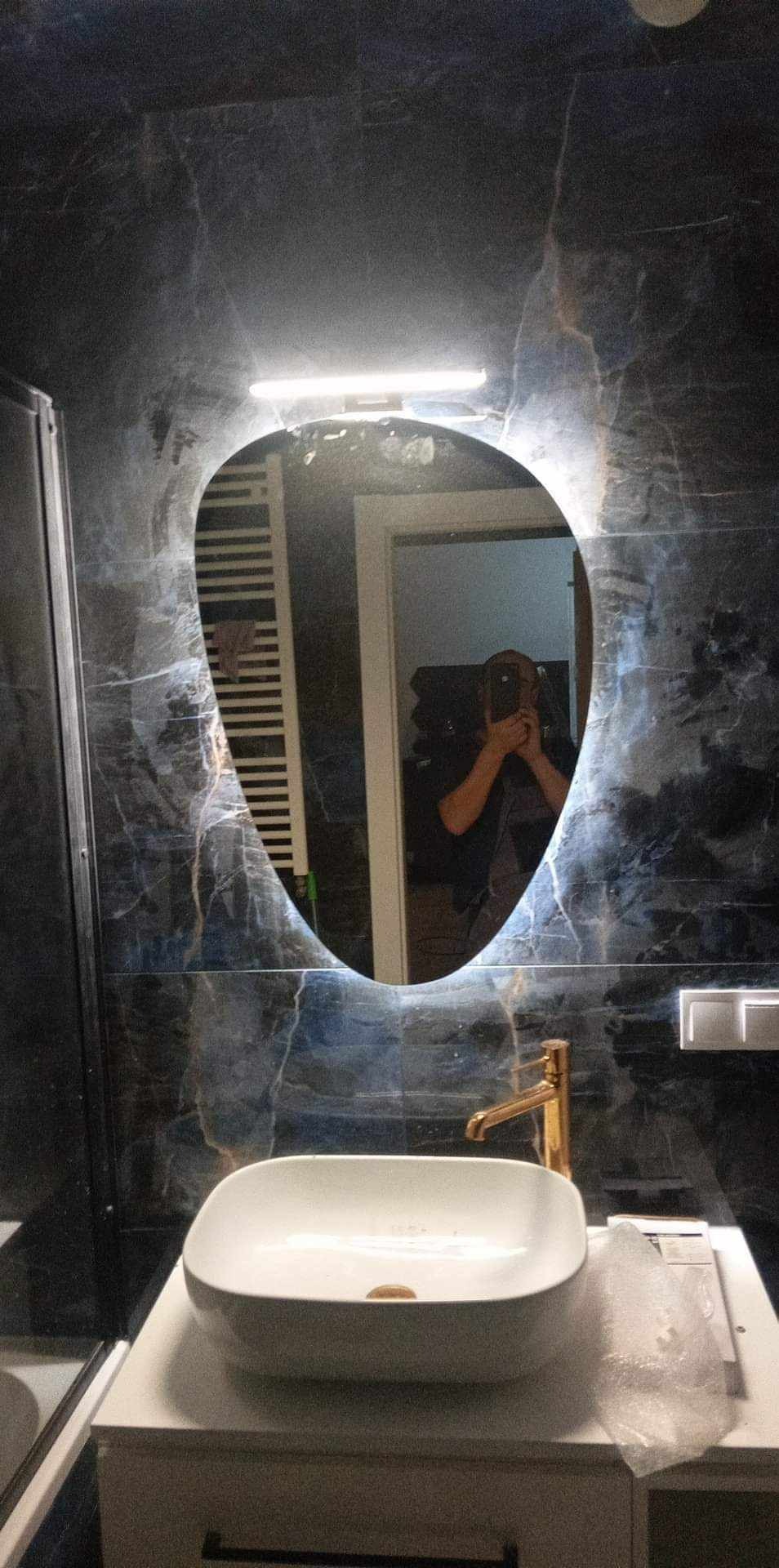 Nowoczesna łazienka z umywalką nablatową, złotą baterią i asymetrycznym lustrem podświetlanym LED, odbicie osoby robiącej zdjęcie.