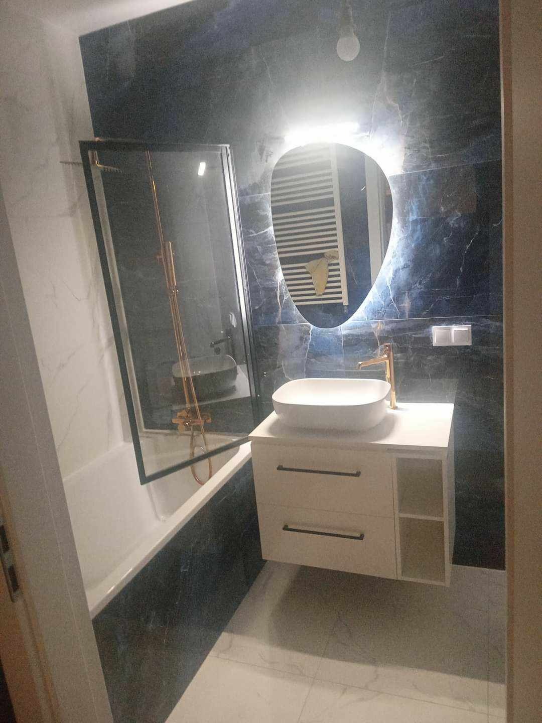 Nowoczesna łazienka z czarnymi marmurowymi płytkami, owalnym lustrem z podświetleniem LED, białą umywalką nablatową i złotą armaturą, widoczna kabina prysznicowa z czarnymi ramami.