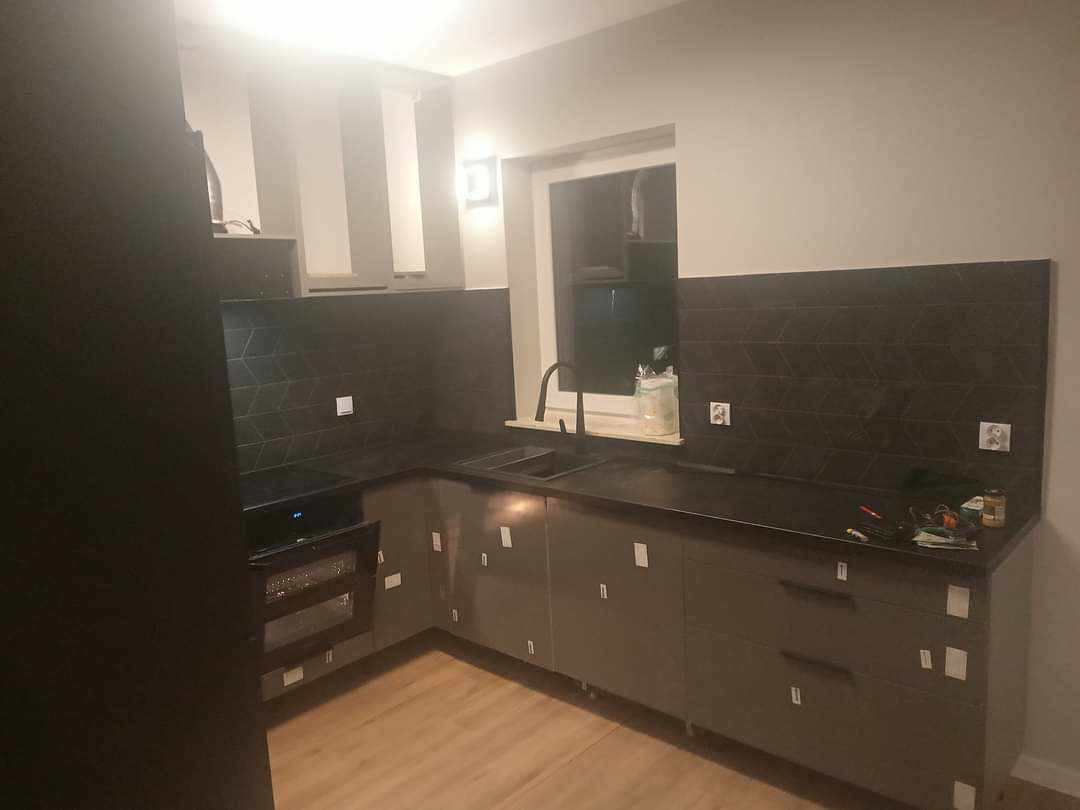 Nowoczesna kuchnia w trakcie montażu: szare szafki z folią ochronną, czarny blat i płytki z geometrycznym wzorem w jodełkę na ścianie, widoczne narzędzia na blacie, jasne drewnopodobne panele...