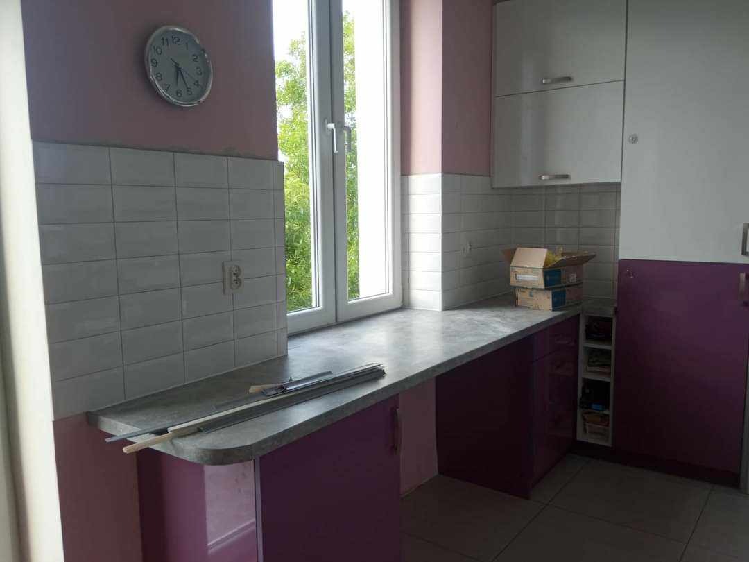 Fragment kuchni w trakcie remontu: szare płytki nad blatem, fioletowe szafki, okno z widokiem na zieleń, widoczne narzędzia i materiały budowlane na blacie, oraz kartony na szafce.