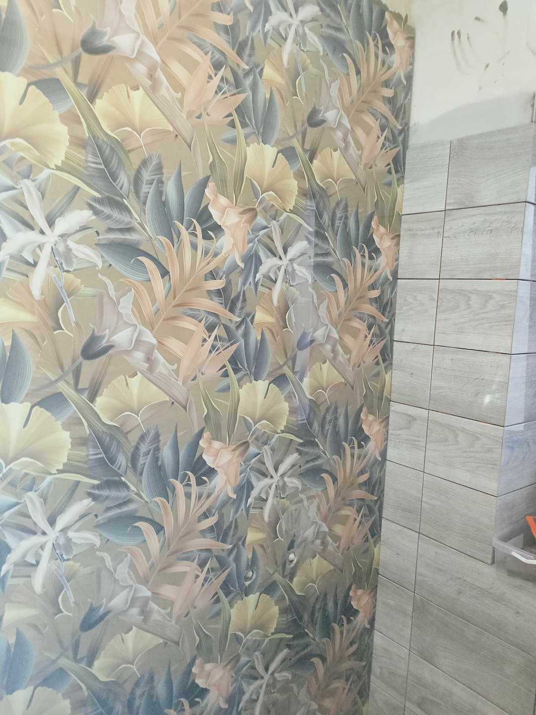 Ściana z wzorzystą tapetą w motywy roślinne sąsiadująca z ułożonymi szarymi płytkami ceramicznymi w trakcie prac wykończeniowych.