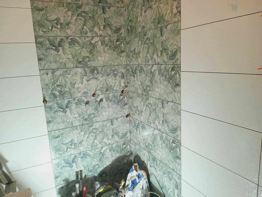 Wnętrze łazienki w trakcie wykańczania: ściana z poziomych, białych płytek sąsiaduje ze ścianą wyłożoną płytkami z motywem liści, widoczne pomarańczowe krzyżyki dystansowe i worek z zaprawą klejową.