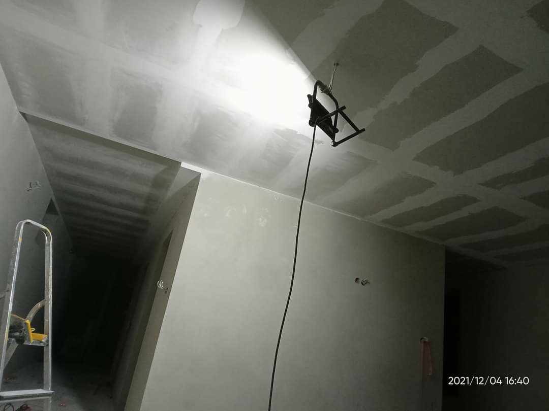 Świeżo położona gładź na suficie podświetlona lampą budowlaną, widoczne ślady szlifowania, drabina aluminiowa w rogu pomieszczenia.