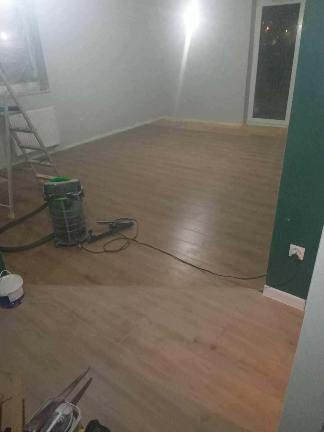Pokój po ułożeniu paneli podłogowych, widoczny odkurzacz przemysłowy, drabina malarska i częściowo pomalowana ściana na zielono.