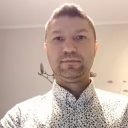 Portret mężczyzny w kwiecistej koszuli, na ścianie dekoracyjny wieszak na ubrania i naklejki w kształcie liści, oświetlenie sufitowe.