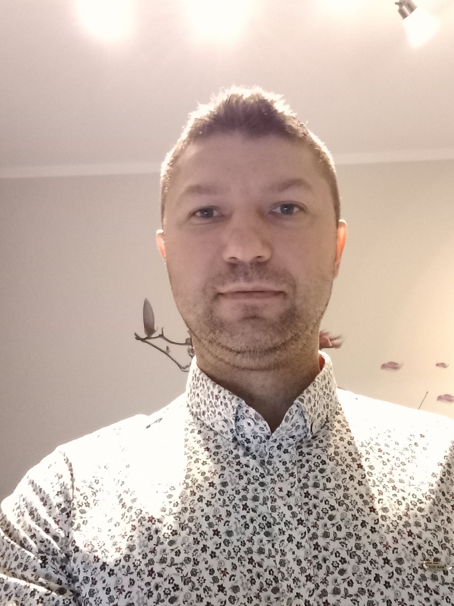 Portret mężczyzny w kwiecistej koszuli, na ścianie dekoracyjny wieszak na ubrania i naklejki w kształcie liści, oświetlenie sufitowe.