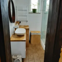 Nowoczesna łazienka z drewnianymi akcentami: blat z jasnego drewna pod umywalką nablatową, okrągłe lustro w ciemnej ramie, drewniana podłoga imitująca panele, kosz na pranie z napisem 'Lights...