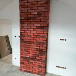 Wykończony fragment ściany z cegły klinkierowej w surowym wnętrzu, widoczne gniazdka elektryczne bez osprzętu.