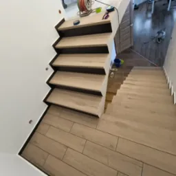 Jasne panele imitujące drewno na podłodze i schodach, widoczne narzędzia i akcesoria na górnym podeście, białe ściany, perspektywa z góry.