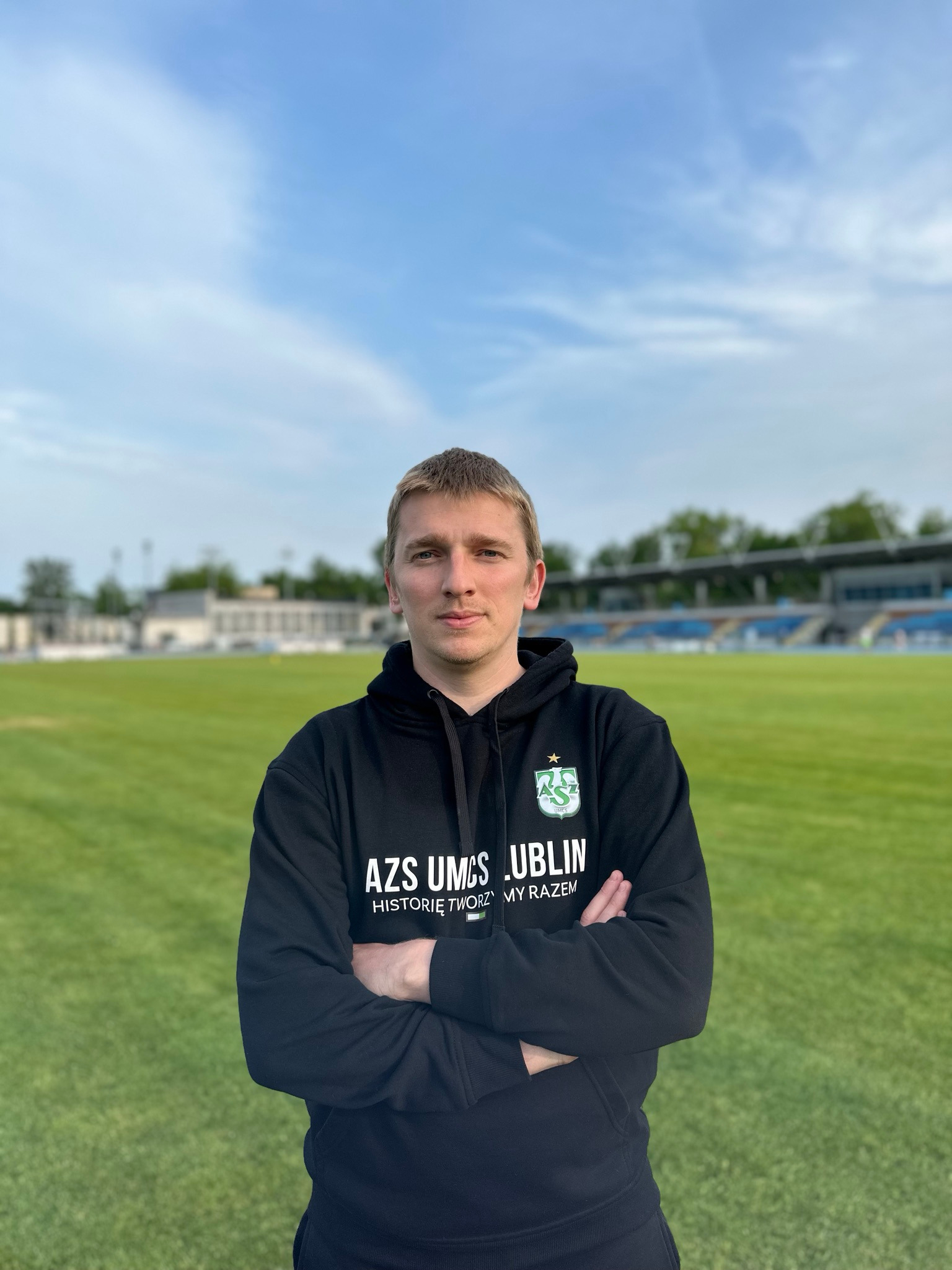 Młody mężczyzna w czarnej bluzie z logo AZS UMCS Lublin na tle stadionu i błękitnego nieba. Skrzyżowane ręce, stonowany wyraz twarzy, naturalne światło.