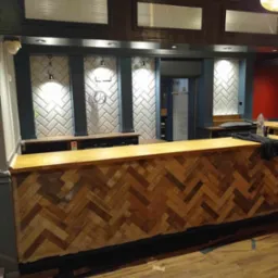 Bar z drewnianym blatem ułożonym w jodełkę, podświetlona ściana z białych płytek w kształcie cegiełek, kolumny w kolorze granatowym, widoczne elementy instalacji elektrycznej.