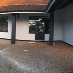 Wykończona elewacja budynku z pomalowanymi na czarno kolumnami i stolarką okienną, teren wokół w trakcie przygotowania, widoczny gruz i worki budowlane.