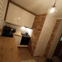 Nowoczesna kuchnia z białymi, błyszczącymi szafkami, drewnianym blatem i ścianą z cegły, oświetlona geometryczną lampą wiszącą. Na blacie widoczny czarny czajnik.