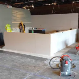 Konstrukcja ścianki działowej z płyt kartonowo-gipsowych w trakcie montażu; widoczny odkurzacz przemysłowy, poziomica, ubranie robocze oraz taśmy ochronne na podłodze.