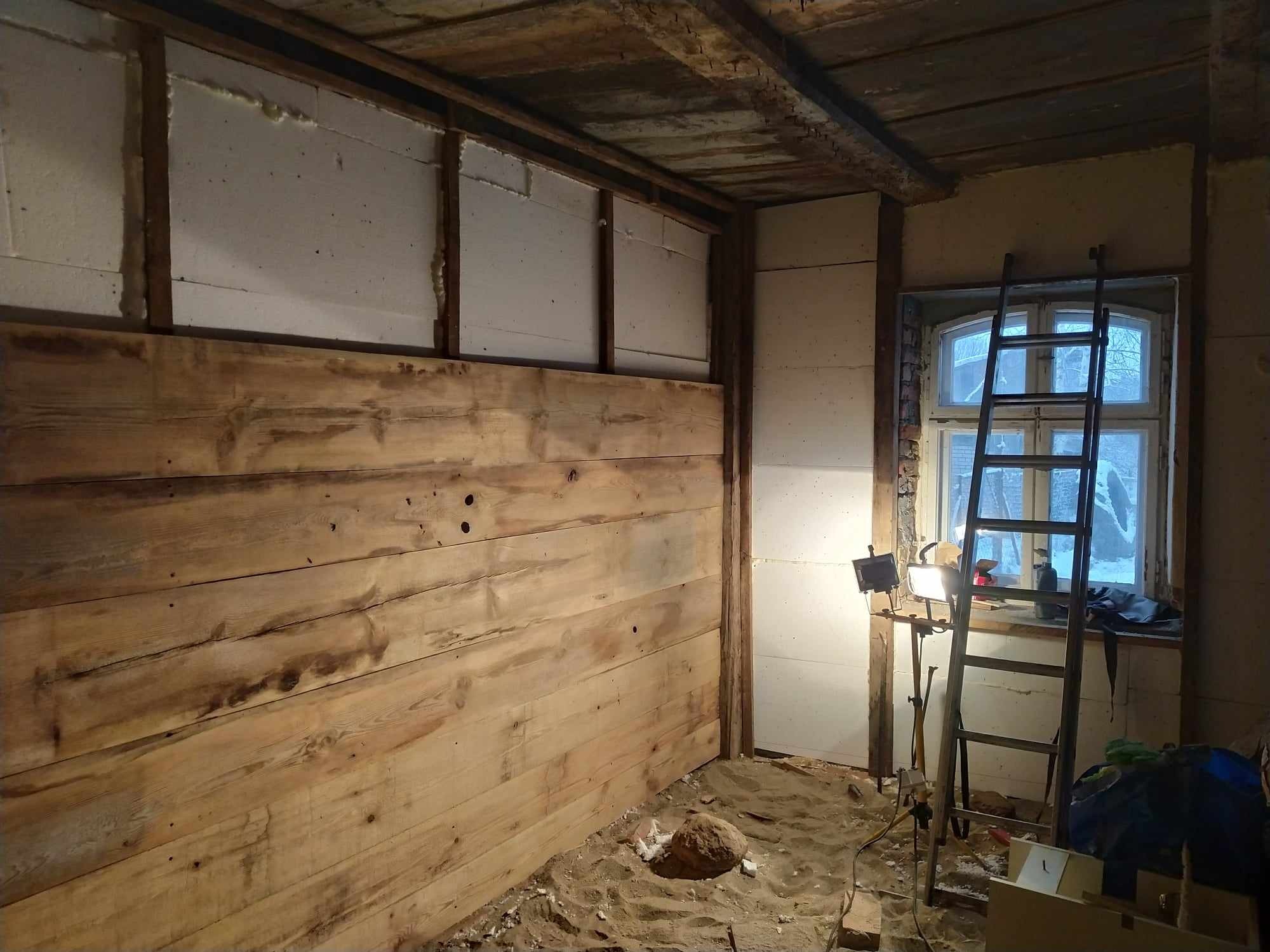 Wnętrze remontowanego pokoju z ocieplonymi ścianami styropianem i częściowo wyłożonymi surowym drewnem, drabina oparta o okno, lampa budowlana na statywie, piasek na podłodze.