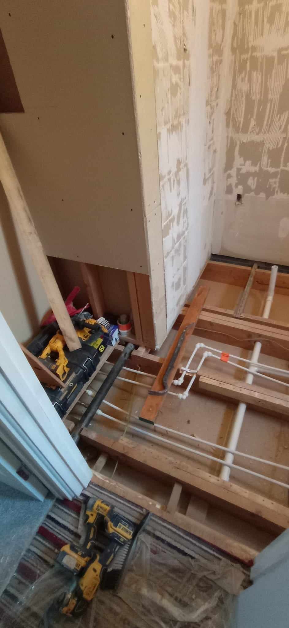 Wnętrze łazienki w trakcie demontażu: odsłonięte rury wodne w podłodze, ściany częściowo bez płytek, narzędzia DeWalt i materiały budowlane w rogu pomieszczenia.