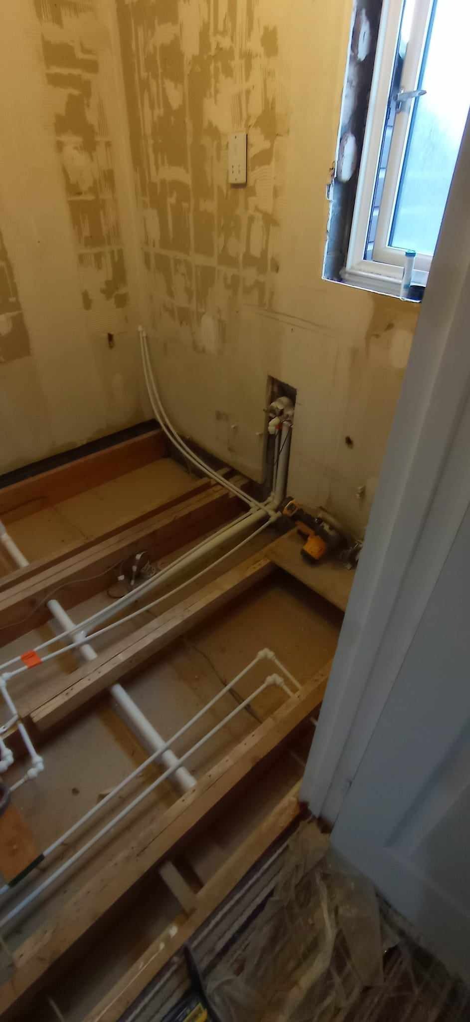 Łazienka w trakcie remontu: widoczne odsłonięte deski podłogowe, instalacja wodna z białych rur, fragmenty starej farby na ścianach i okno.