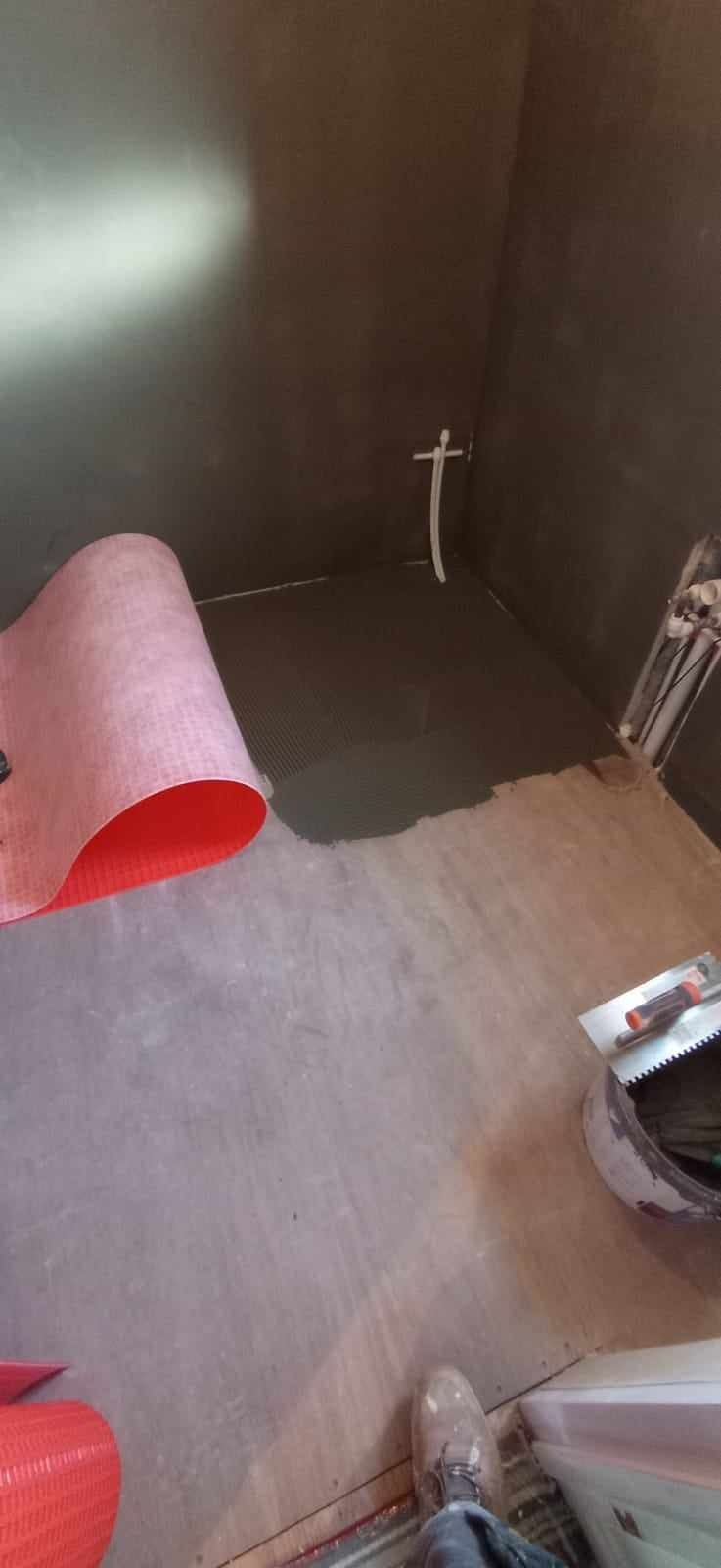 Przygotowanie podłoża pod płytki w łazience: rozłożona czerwona mata izolacyjna na wylewce, widoczna warstwa zaprawy klejowej na podłodze, rury instalacyjne w ścianie, narzędzia glazurnicze...