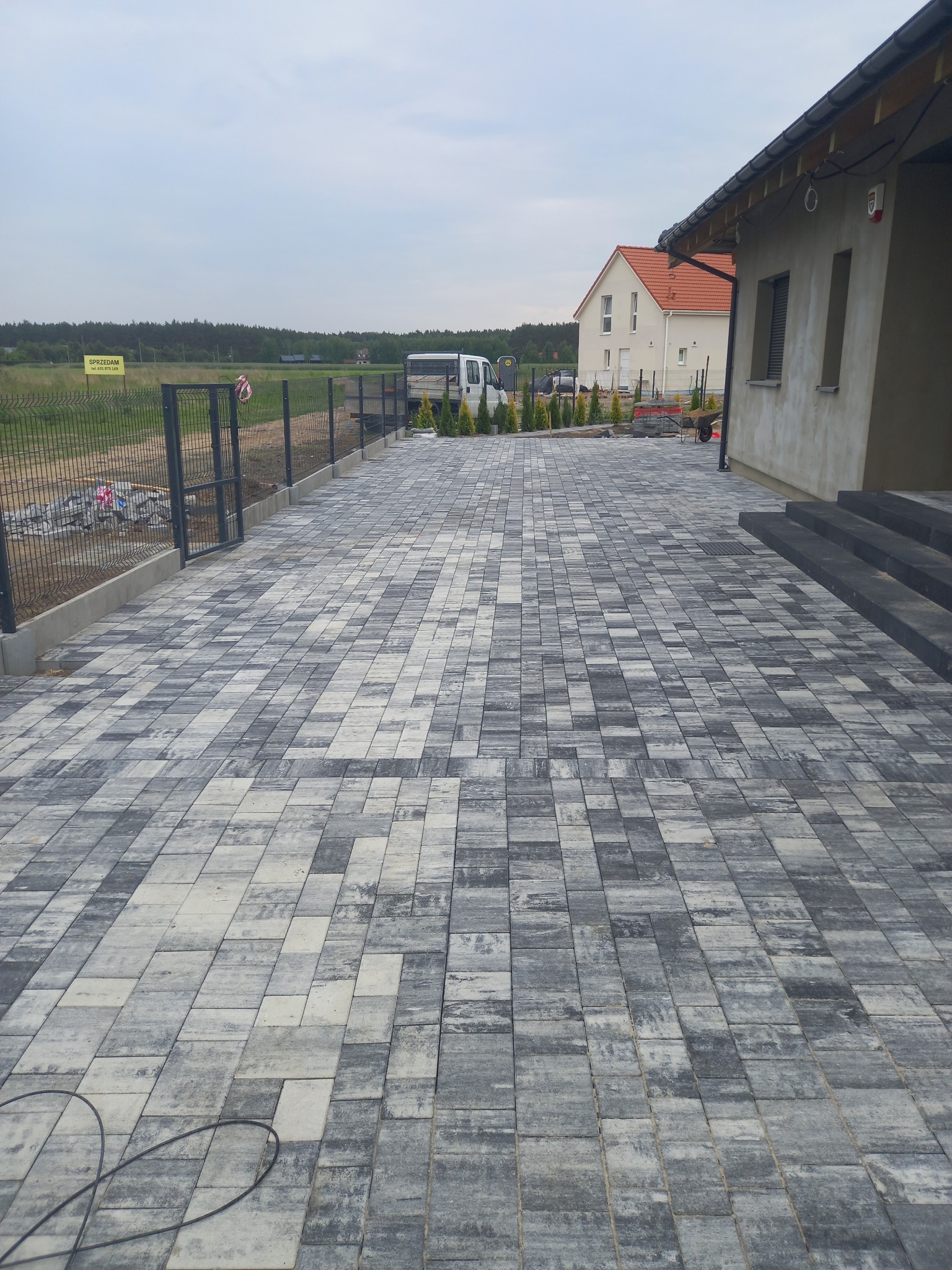 Nowo ułożony, wzorzysty podjazd z kostki brukowej w odcieniach szarości, prowadzący do domu z widocznymi schodami wejściowymi i ogrodzeniem panelowym w tle.