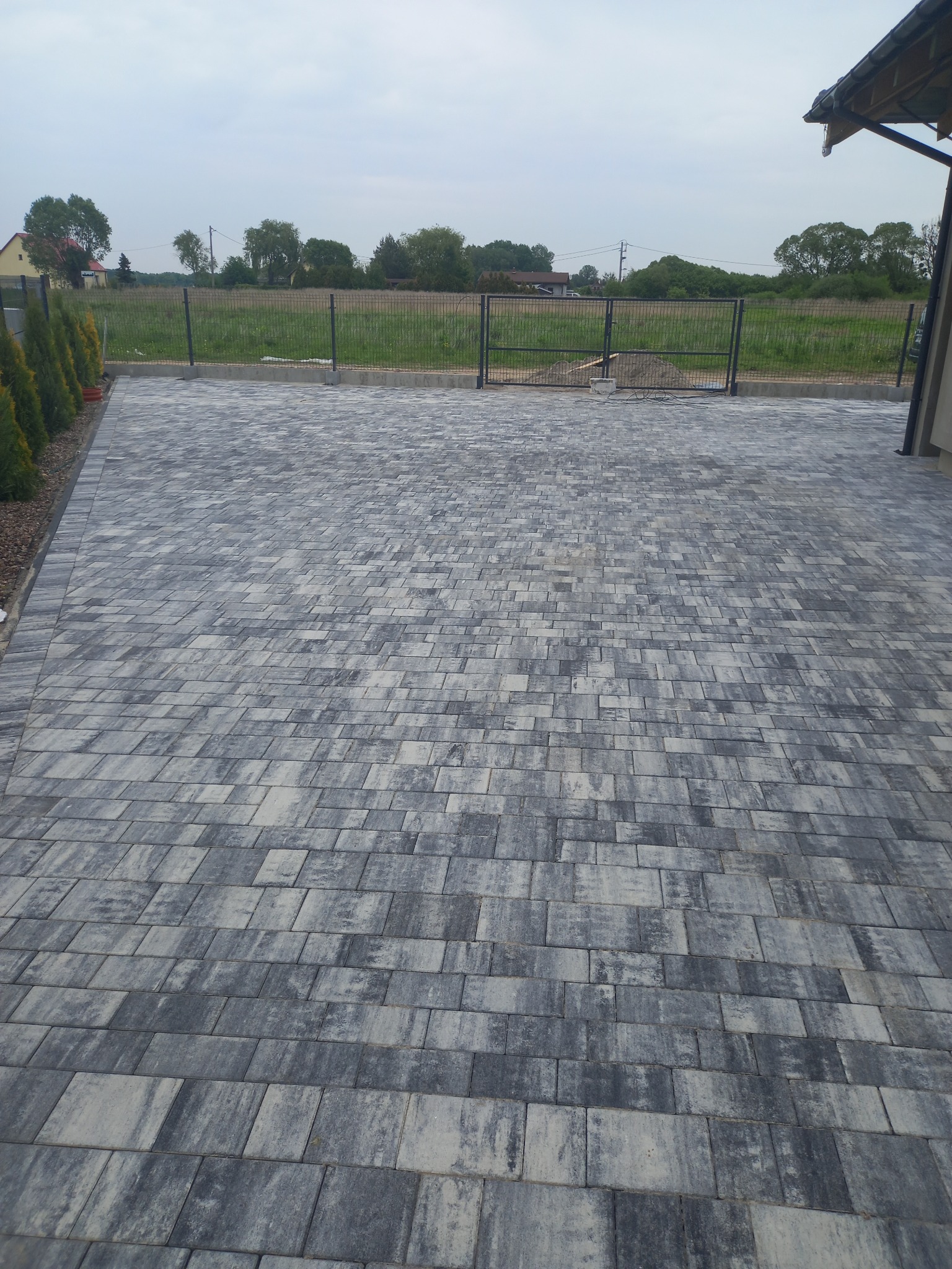 Ułożona szara kostka brukowa na podjeździe, widok na ogrodzenie panelowe i zieleń w tle.