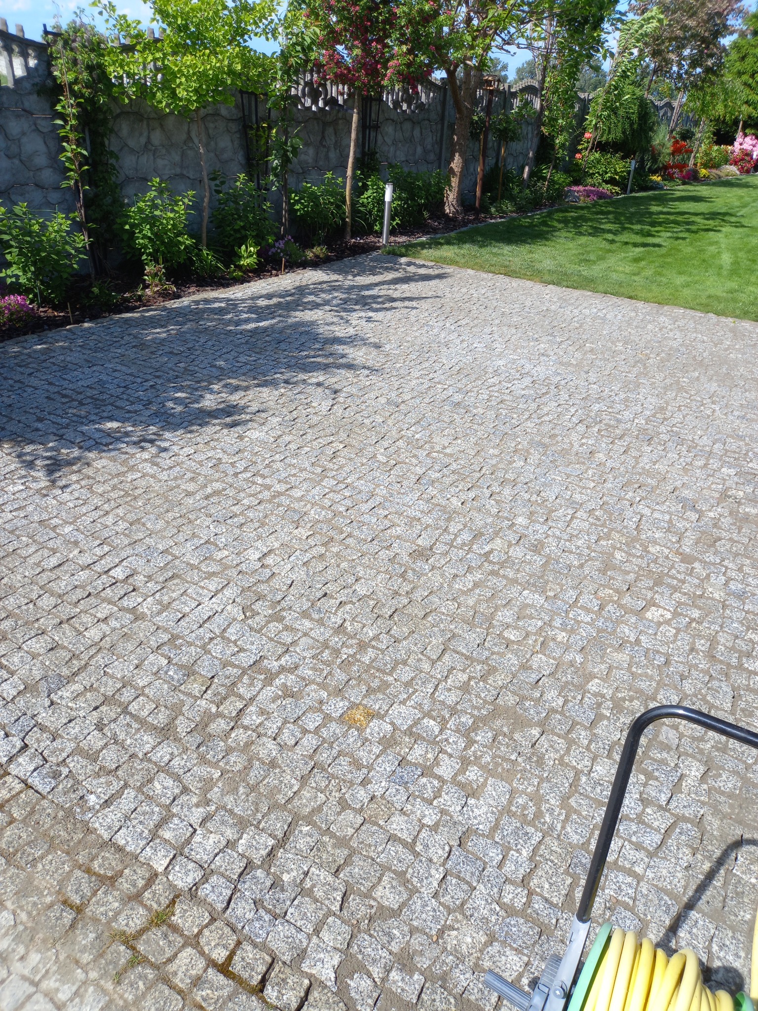 Wyłożona kostką granitową nawierzchnia w Pyzdrach, otoczona zielenią i ozdobnym ogrodzeniem, z widocznym w rogu wózkiem na wąż ogrodowy.