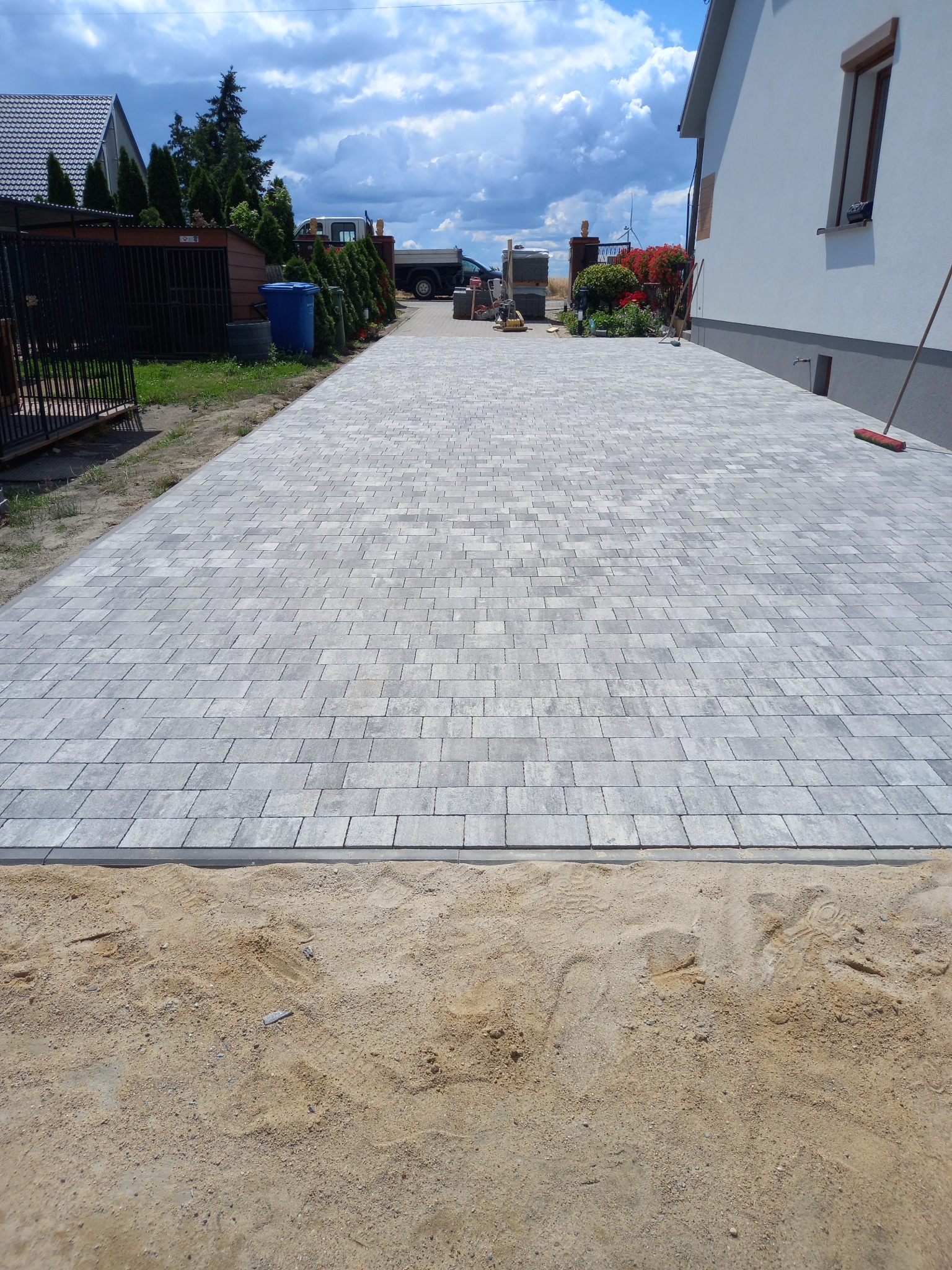 Szeroki podjazd z szarej kostki brukowej ułożonej we wzór jodełki, z widocznymi śladami opon na piasku po bokach, prowadzący do domu z ogrodem i zaparkowaną ciężarówką w tle.