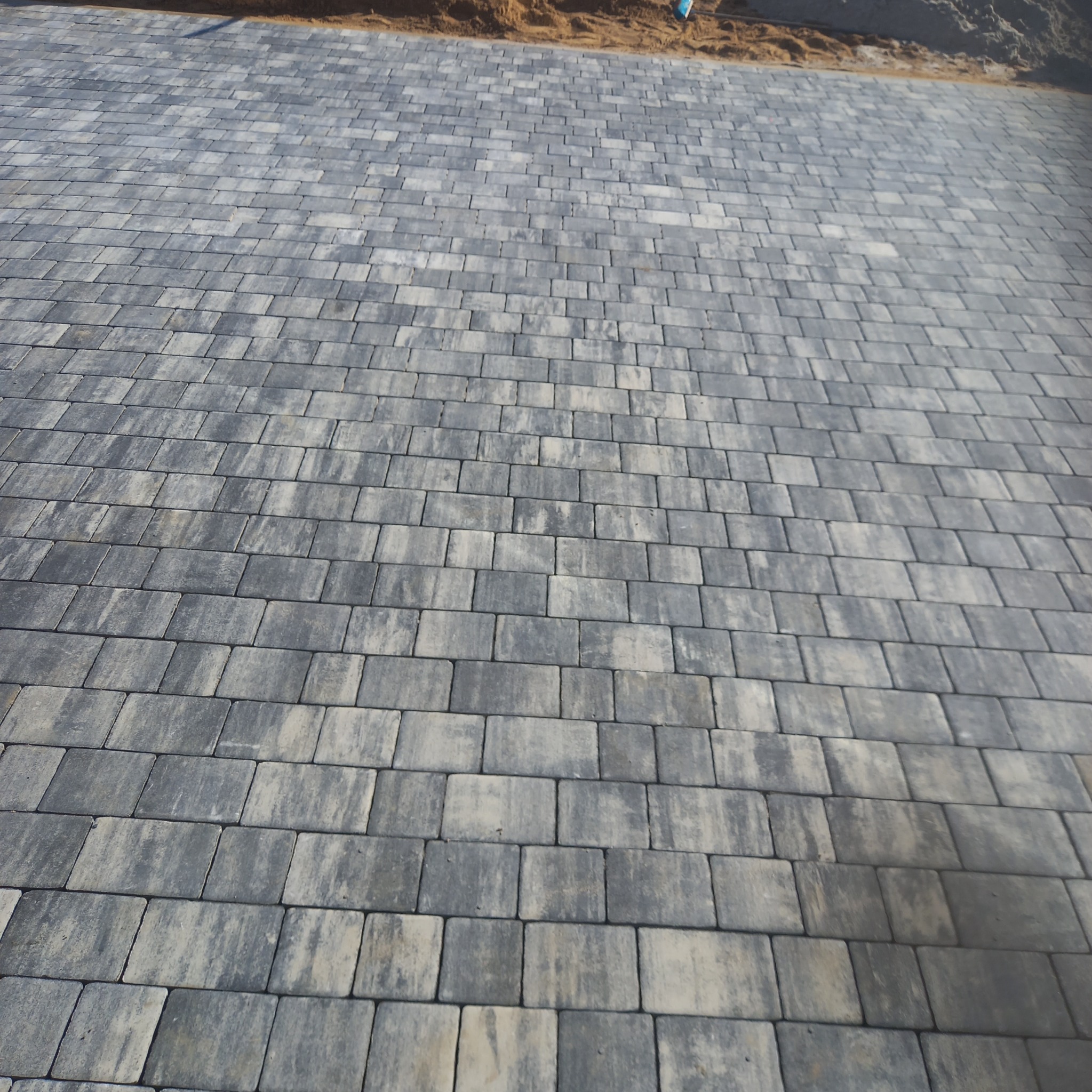 Ułożona szara kostka brukowa o prostokątnym kształcie, widziana pod kątem, z piaskiem widocznym w tle.