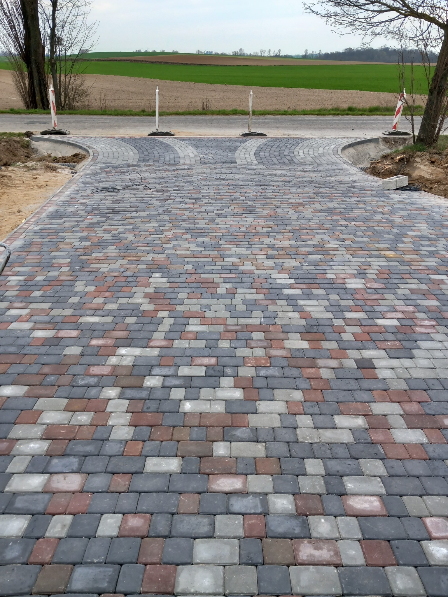 Szeroki, wybrukowany podjazd z kostki w odcieniach szarości, czerwieni i bieli, rozwidlający się na dwa kierunki, z widocznymi słupkami ostrzegawczymi i polami w tle.