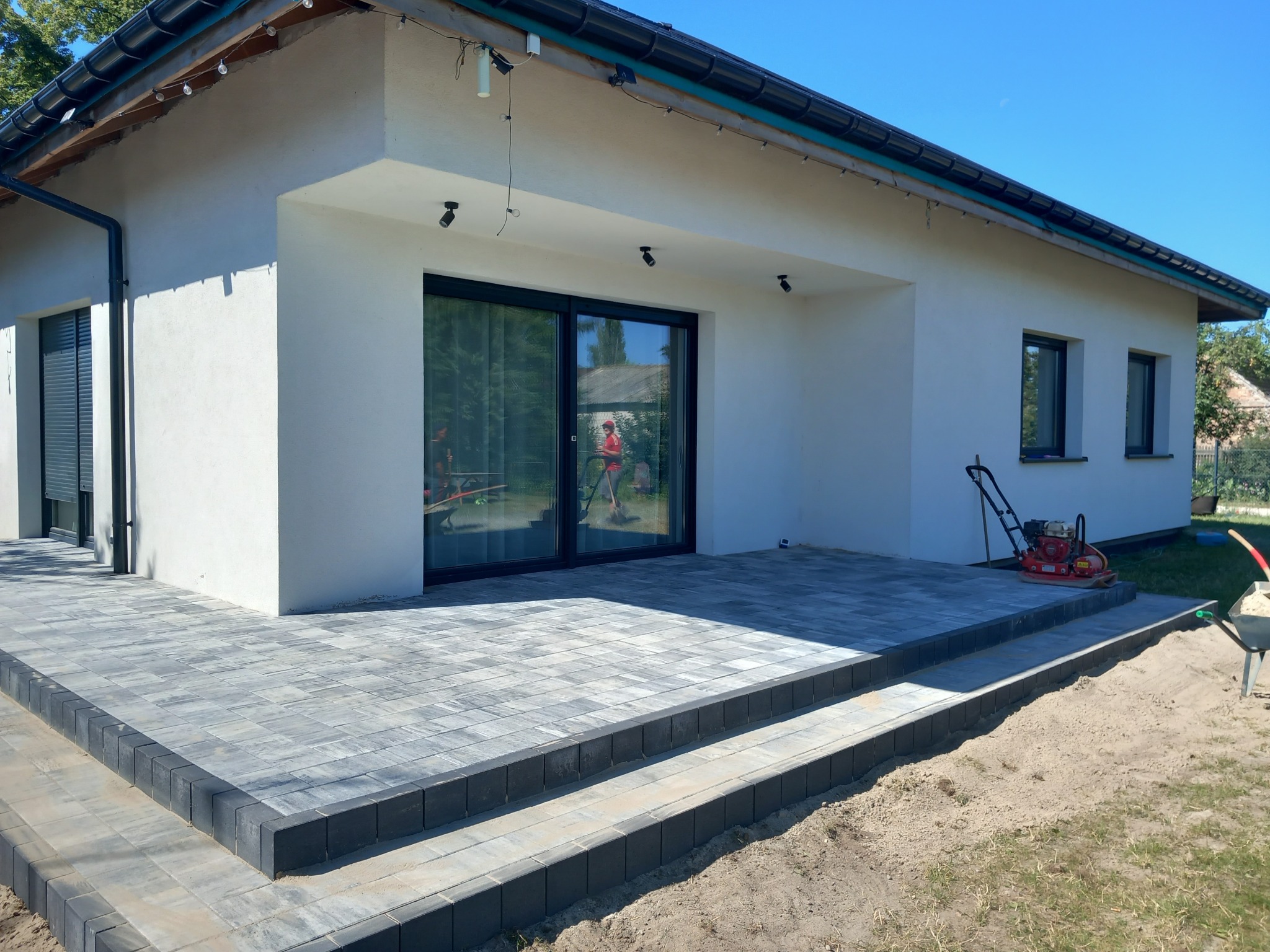 Nowoczesny taras z szarej kostki brukowej ułożonej w geometryczny wzór, z dwoma stopniami obłożonymi ciemnoszarą kostką, przylegający do białego domu z dużymi przesuwnymi drzwiami tarasowymi...