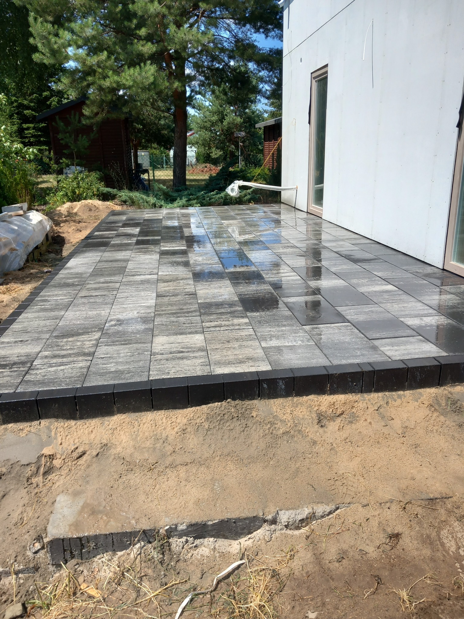Świeżo ułożony taras z kostki brukowej w odcieniach szarości, obramowany ciemnymi obrzeżami, przylegający do białego budynku z widocznym oknem, otoczony piaskiem i zielenią.
